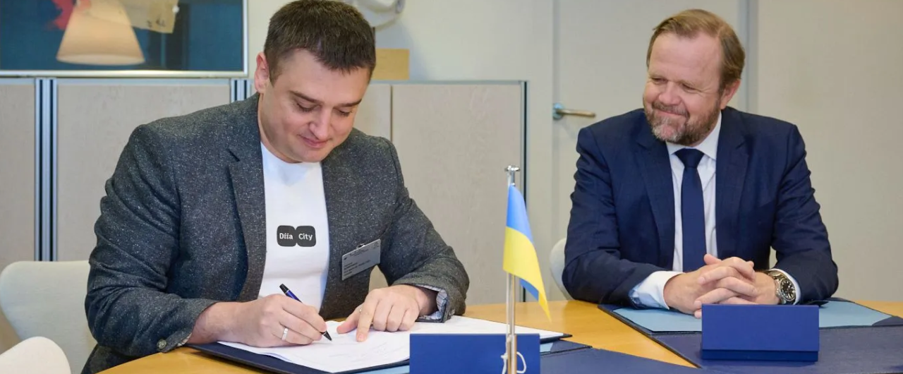 Двоє чоловіків за столом, один підписує документ із українським прапором, інший у костюмі спостерігає, офісне приміщення.